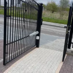 Otwarta, czarna brama wjazdowa z automatyką, widoczny siłownik i fotokomórka, kostka brukowa na podjeździe, skrzynka na listy na drugim skrzydle bramy.