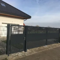 Nowoczesna, przesuwna brama wjazdowa w kolorze grafitowym, z perforowanym wypełnieniem, zamontowana przed domem z jasną elewacją i ciemnym dachem.