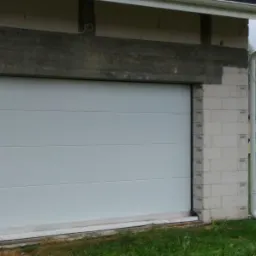 Biała brama garażowa segmentowa zamontowana w surowym stanie budynku z bloczków betonowych, widoczne elementy konstrukcyjne i niezakończone prace wykończeniowe.