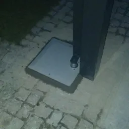 Słupkowa obudowa domofonu lub wideofonu, zamontowana na betonowej podstawie, osadzona w kostce brukowej. Widoczne elementy sterujące na słupku.