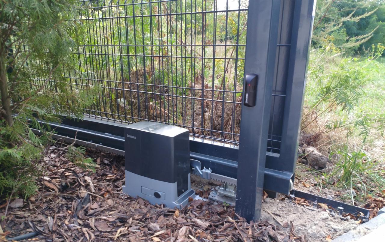 Automat do bramy przesuwnej marki Beninca, zamontowany na szarej bramie z siatki, widoczny czytnik zbliżeniowy, otoczenie zarośnięte trawą i krzewami, ziemia pokryta liśćmi.