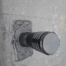 Czarny komin wentylacyjny zamontowany na bitumicznym dachu płaskim, widoczne śruby mocujące i poziomica na górnej części rury.