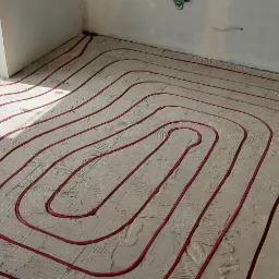 Układ ogrzewania podłogowego z czerwonymi rurami na szarej posadzce w jasnym pomieszczeniu z dużym oknem balkonowym. Widoczne fragmenty ścian i instalacji.