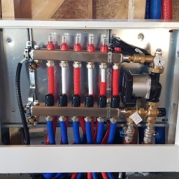 INSTALMIX SP. Z O.O. - Rozdzielacz ogrzewania podłogowego w białej obudowie, z widocznymi rurami w kolorach czerwonym i niebieskim, zamontowany na drewnianej ścianie w surowym stanie.