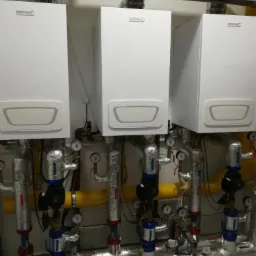 Trzy identyczne, białe kotły grzewcze zamontowane na ścianie, z widocznymi rurami i manometrami, zabezpieczonymi folią izolacyjną.