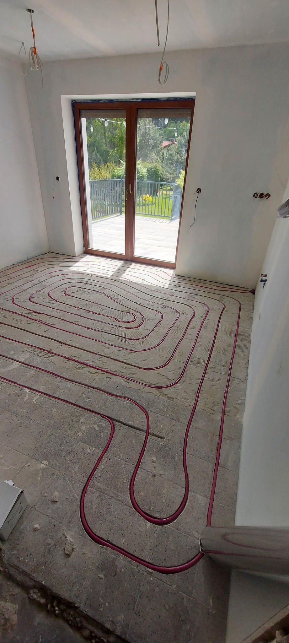 Instalacja ogrzewania podłogowego z rurami w kolorze czerwonym, ułożonymi spiralnie na szarym podłożu w jasnym pomieszczeniu z oknem balkonowym.