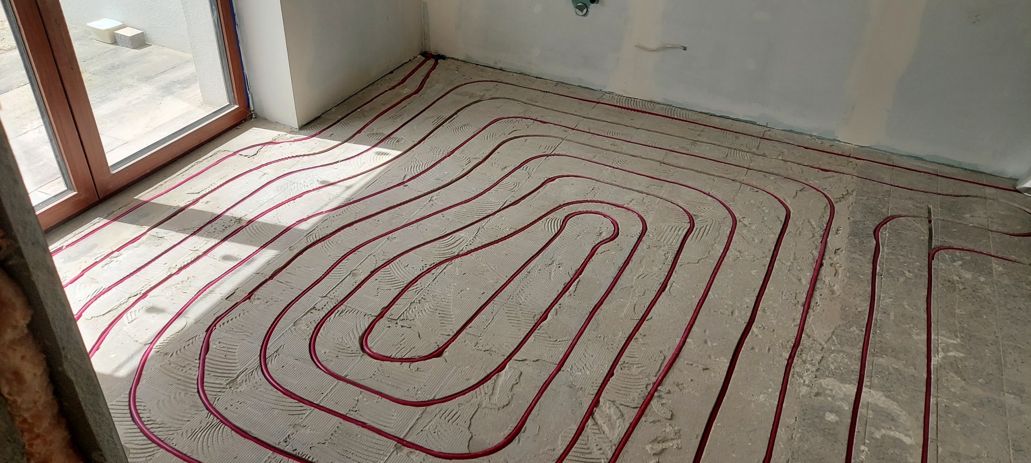 Ułożone, czerwone rury ogrzewania podłogowego na szarej wylewce w jasnym pomieszczeniu z oknem. Widoczne fragmenty ścian i instalacji. Układ spiralny rur.