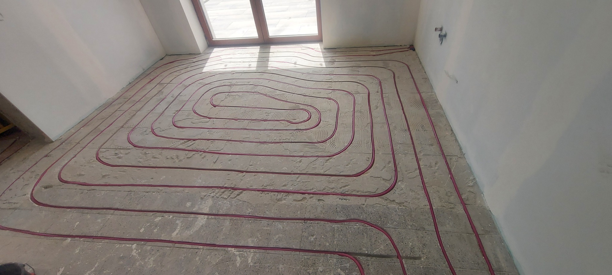 Różowe rury ogrzewania podłogowego ułożone spiralnie na szarym podłożu, widok z góry, okno w tle. Instalacja w surowym stanie przed wylaniem posadzki.