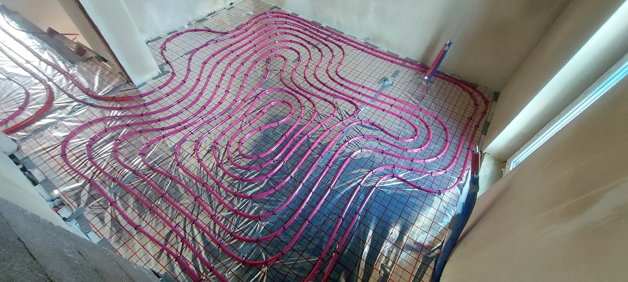 Różowe rury ogrzewania podłogowego ułożone na srebrnej folii z siatką, widok z góry. Częściowo widoczne okno i ściany w surowym stanie.