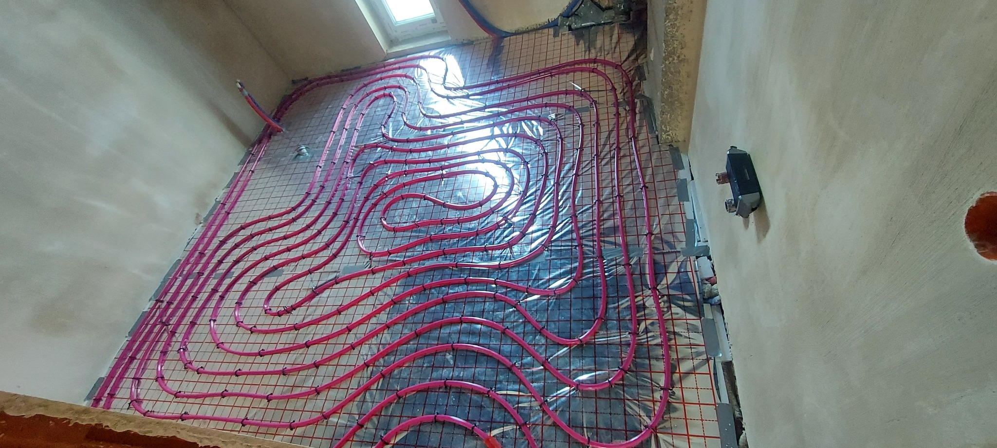 Różowe rury ogrzewania podłogowego ułożone na metalowej siatce w surowym pomieszczeniu z oknem dachowym i grzejnikiem na ścianie. Widok z góry na instalację.