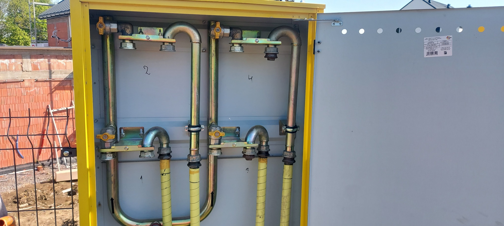 Żółta szafka gazowa z otwartymi drzwiami, widoczne metalowe rury i zawory, tło stanowią elementy budowy.