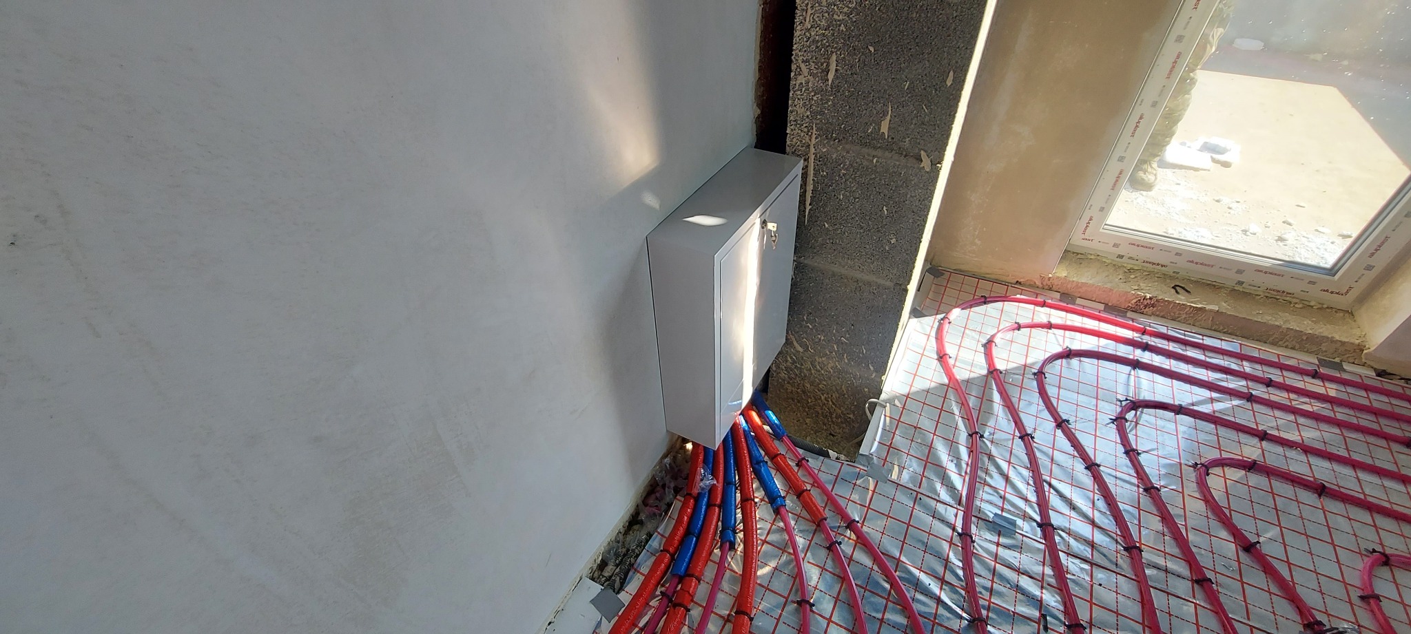 Rozdzielacz ogrzewania podłogowego z rurami w kolorze czerwonym i niebieskim, zamontowany na ścianie w pomieszczeniu z oknem, widok z góry.