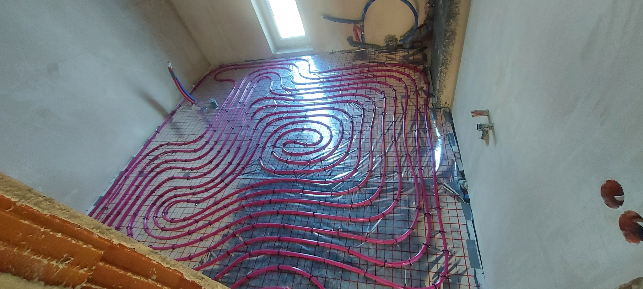 Różowe rury ogrzewania podłogowego ułożone spiralnie na siatce, widok z góry na surową podłogę i ściany w trakcie instalacji, okno dachowe w tle.