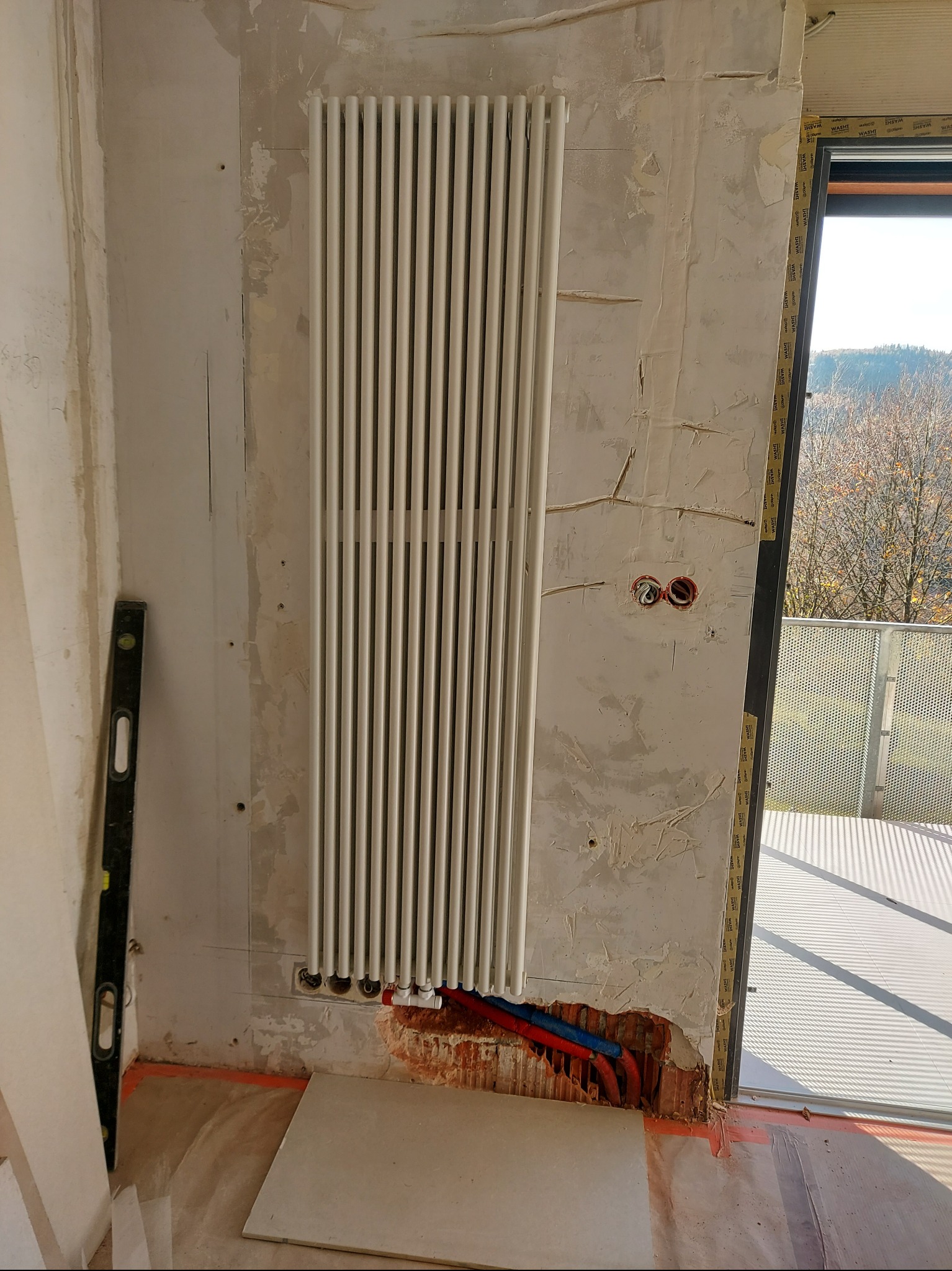 Biały, pionowy grzejnik panelowy zamontowany na surowej ścianie obok okna balkonowego. Widoczne wystające rury instalacji grzewczej i poziomica przy ścianie.