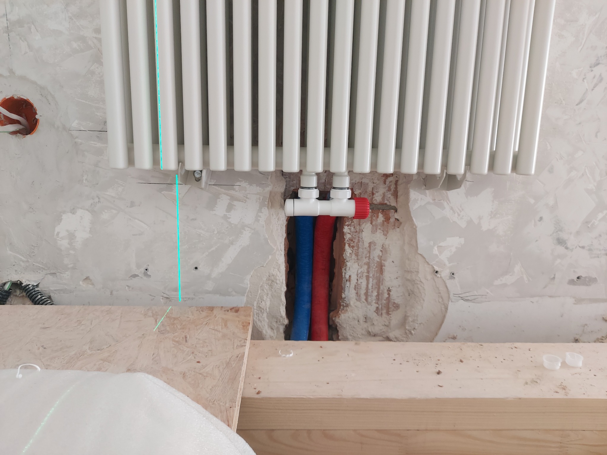 Biały grzejnik żeberkowy na ścianie w trakcie montażu, widoczne niebieskie i czerwone rury instalacji CO oraz fragment lasera krzyżowego.
