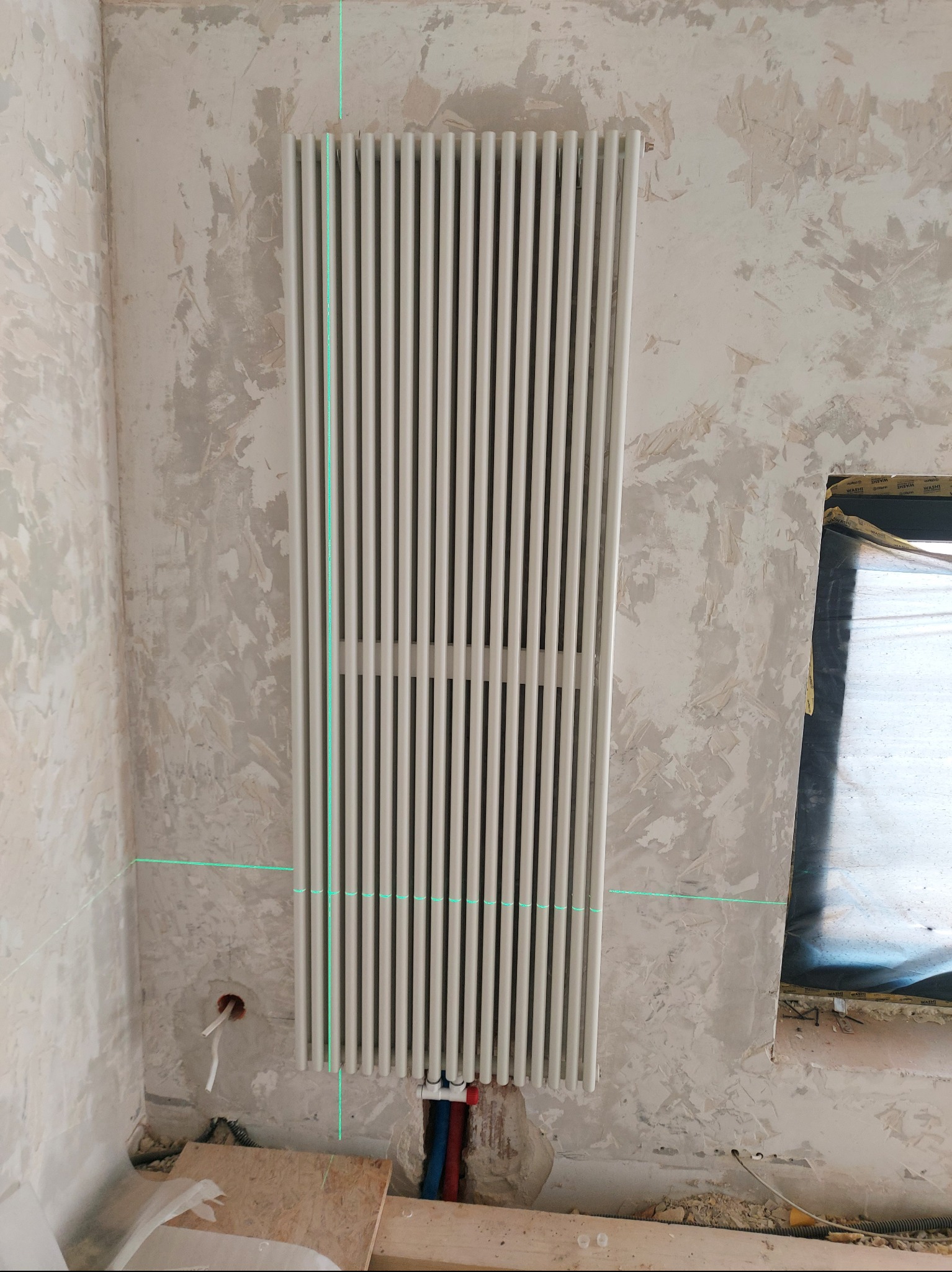 Zamontowany, biały grzejnik żeberkowy na tle surowej ściany z widocznymi przewodami instalacji. Użycie lasera do precyzyjnego montażu.