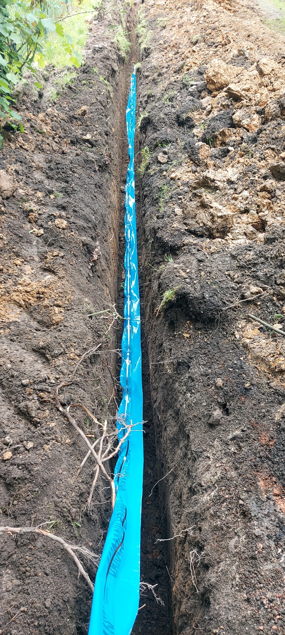 Wąski wykop w ziemi, zabezpieczony niebieską folią z nadrukiem, prawdopodobnie pod instalację wodno-kanalizacyjną. Widoczne ślady po wykopie i korzenie.