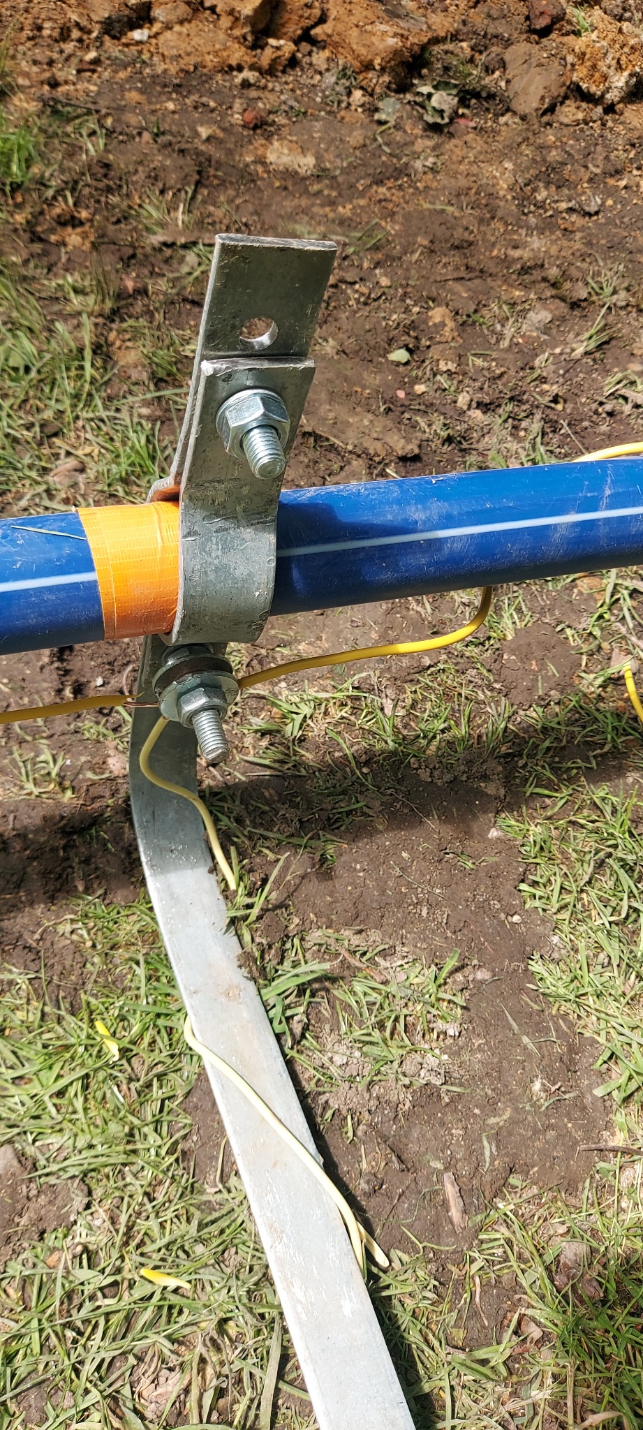 Mocowanie rury gazowej z uziemieniem na zewnątrz, zabezpieczone taśmą. Metalowy wspornik przytwierdzony do gruntu. Instalacja w trawie i ziemi.