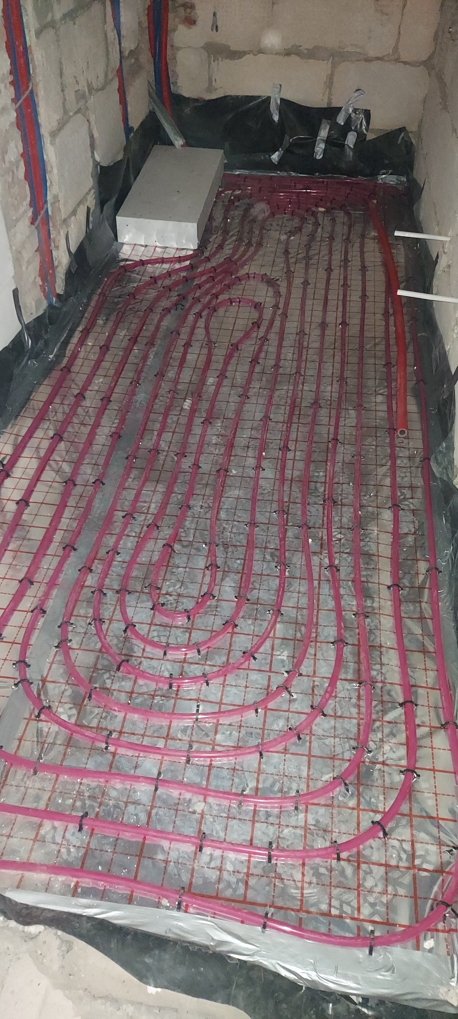 Różowe rury ogrzewania podłogowego zamocowane na siatce, przygotowane do zalania posadzką w Bielsko-Białej. Widoczne izolacja i fragment ściany.