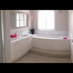 Jasna łazienka z marmurowym blatem, dwiema umywalkami, lustrem z podziałami, wanną zabudowaną panelami, schodkami i kwiatem orchidei.