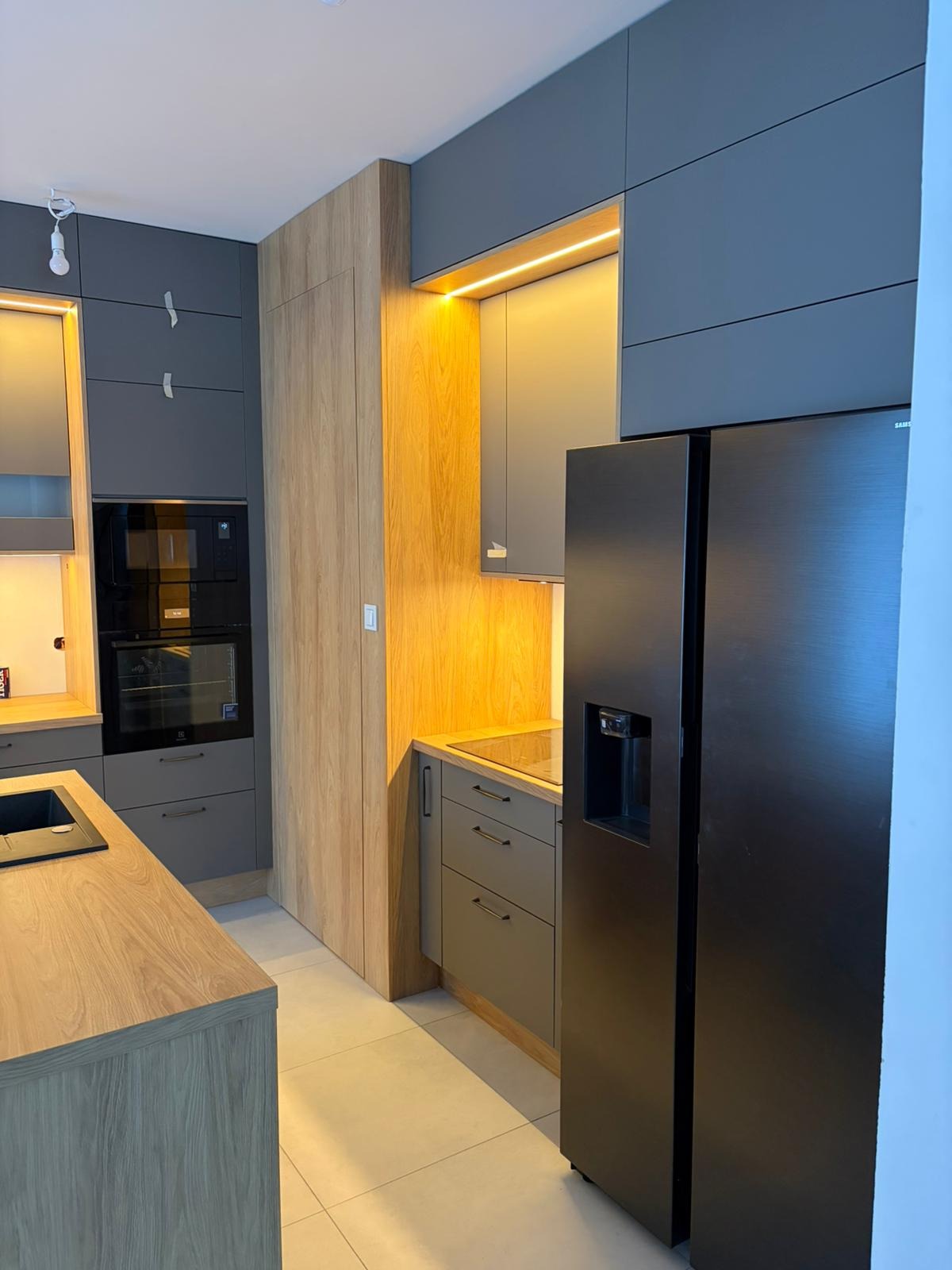 Nowoczesna kuchnia: szare szafki, drewniane akcenty, oświetlenie LED i czarna lodówka side-by-side. Minimalistyczny design z dbałością o detale.