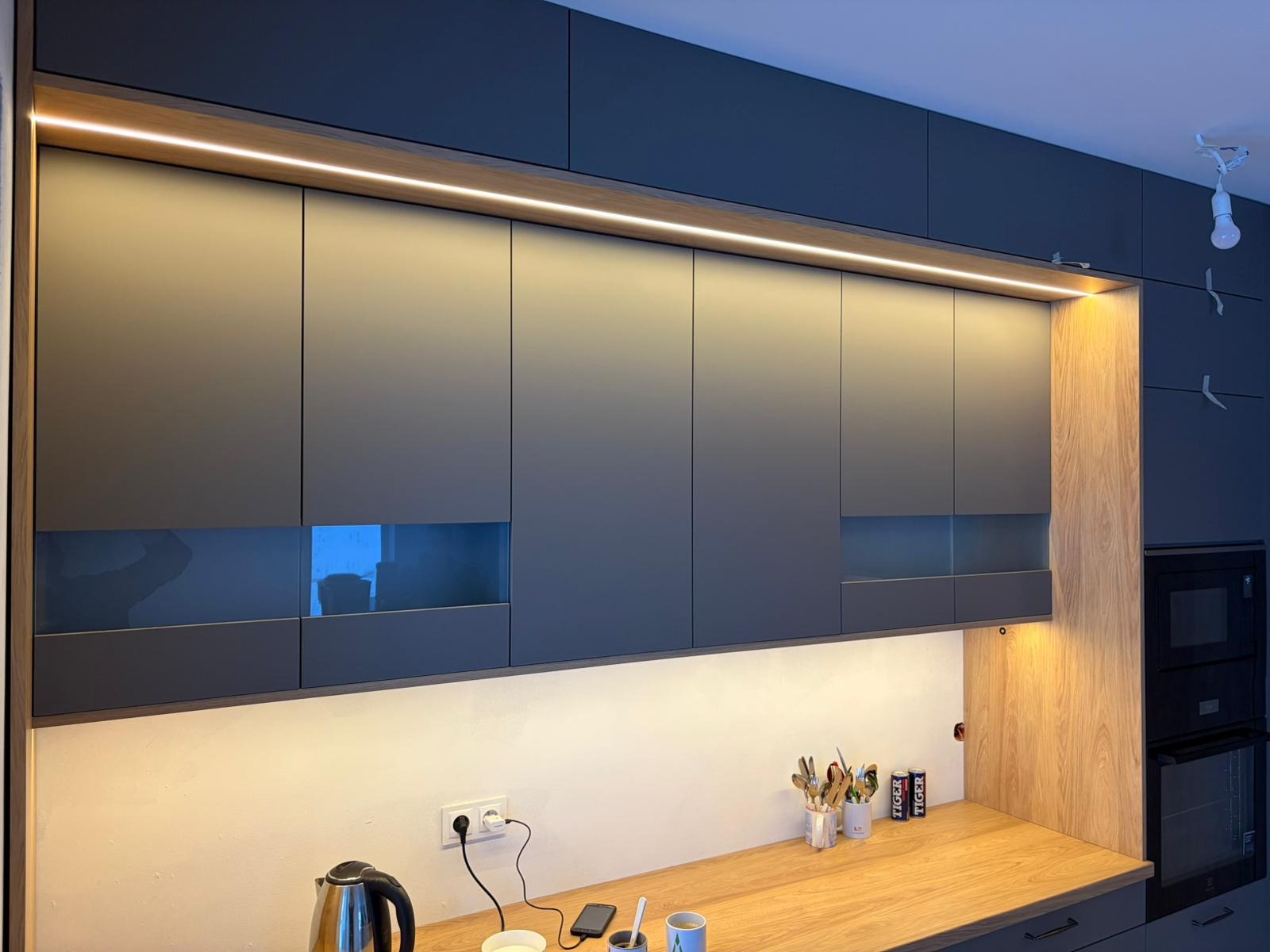 Nowoczesna kuchnia: szare szafki górne z podświetleniem LED, drewniany blat, widoczne akcesoria kuchenne i gniazdko elektryczne. Minimalistyczny design.