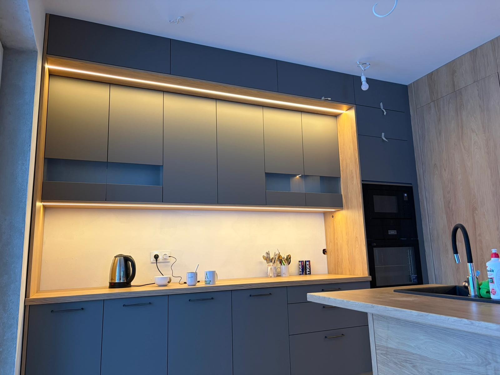 Nowoczesna kuchnia z szarymi szafkami, drewnianym blatem i oświetleniem LED. Widoczne AGD w zabudowie i akcesoria kuchenne. Minimalistyczny design z dbałością o detale.