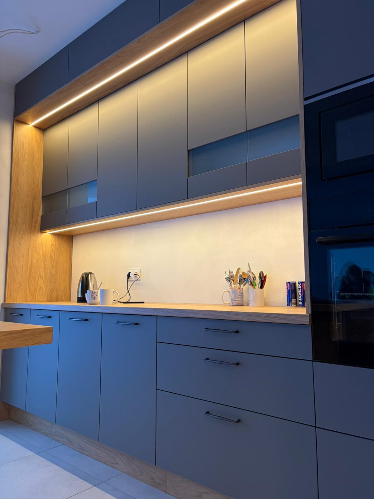 Nowoczesna kuchnia z szarymi frontami, drewnianym blatem i oświetleniem LED. Widoczne szafki górne z przeszkleniami i sprzęt AGD w zabudowie. Minimalistyczny design.