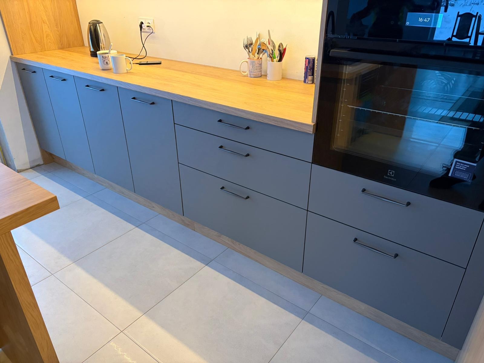 Szare szafki kuchenne z drewnianym blatem i piekarnikiem Electrolux. Minimalistyczny design, widoczne akcesoria kuchenne i fragment stołu. Jasne płytki podłogowe.