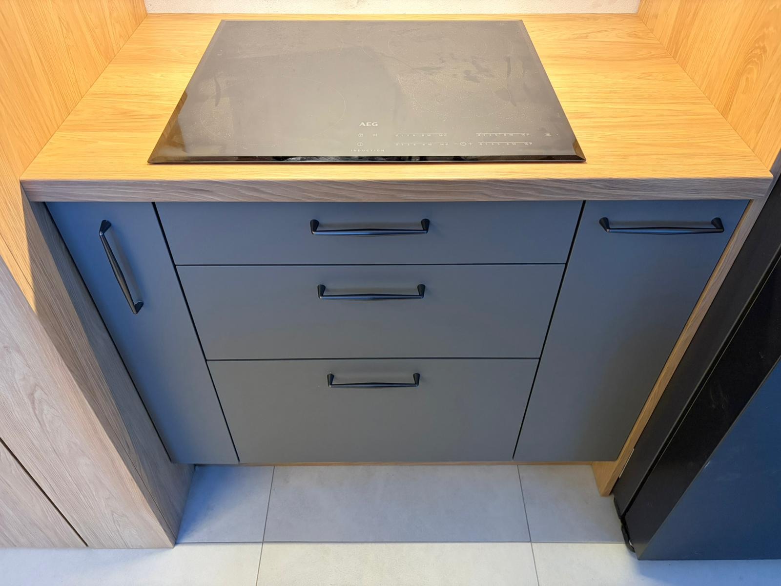 Szare szafki kuchenne z trzema szufladami i drewnianym blatem, na którym zamontowana jest płyta indukcyjna AEG. Minimalistyczny design, jasna podłoga.