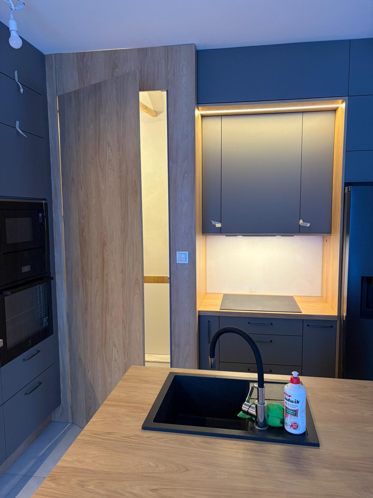 Nowoczesna kuchnia z szarymi szafkami, drewnianym blatem i ukrytymi drzwiami do spiżarni. Oświetlenie LED podkreśla ciepło drewna.