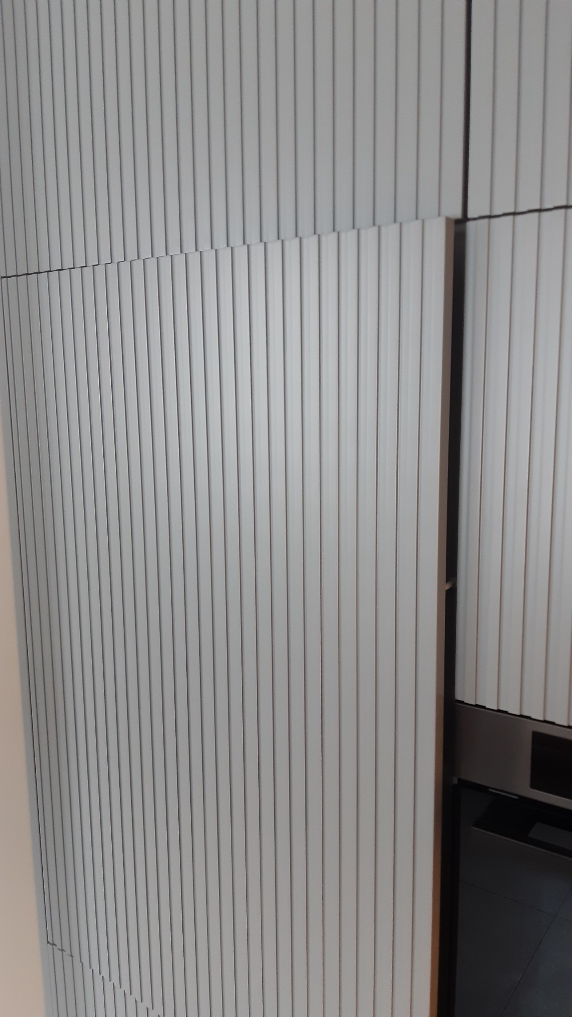 Szara, nowoczesna szafka kuchenna z ryflowanymi frontami, widoczny fragment piekarnika. Minimalistyczny design, pionowe linie, subtelne oświetlenie.