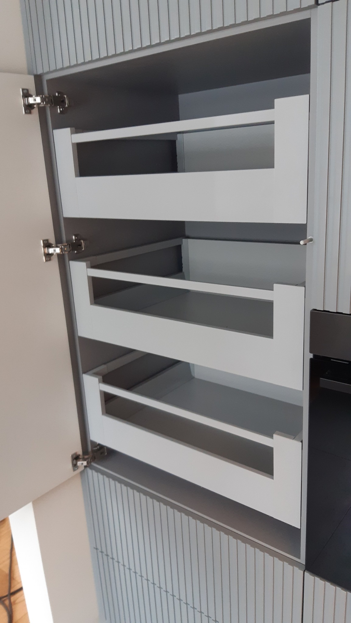 Wnętrze szafki kuchennej z wysuwanymi, metalowymi półkami w odcieniach szarości. Widoczne zawiasy i fragment frontu szafki. Pionowe żłobienia na obudowie.