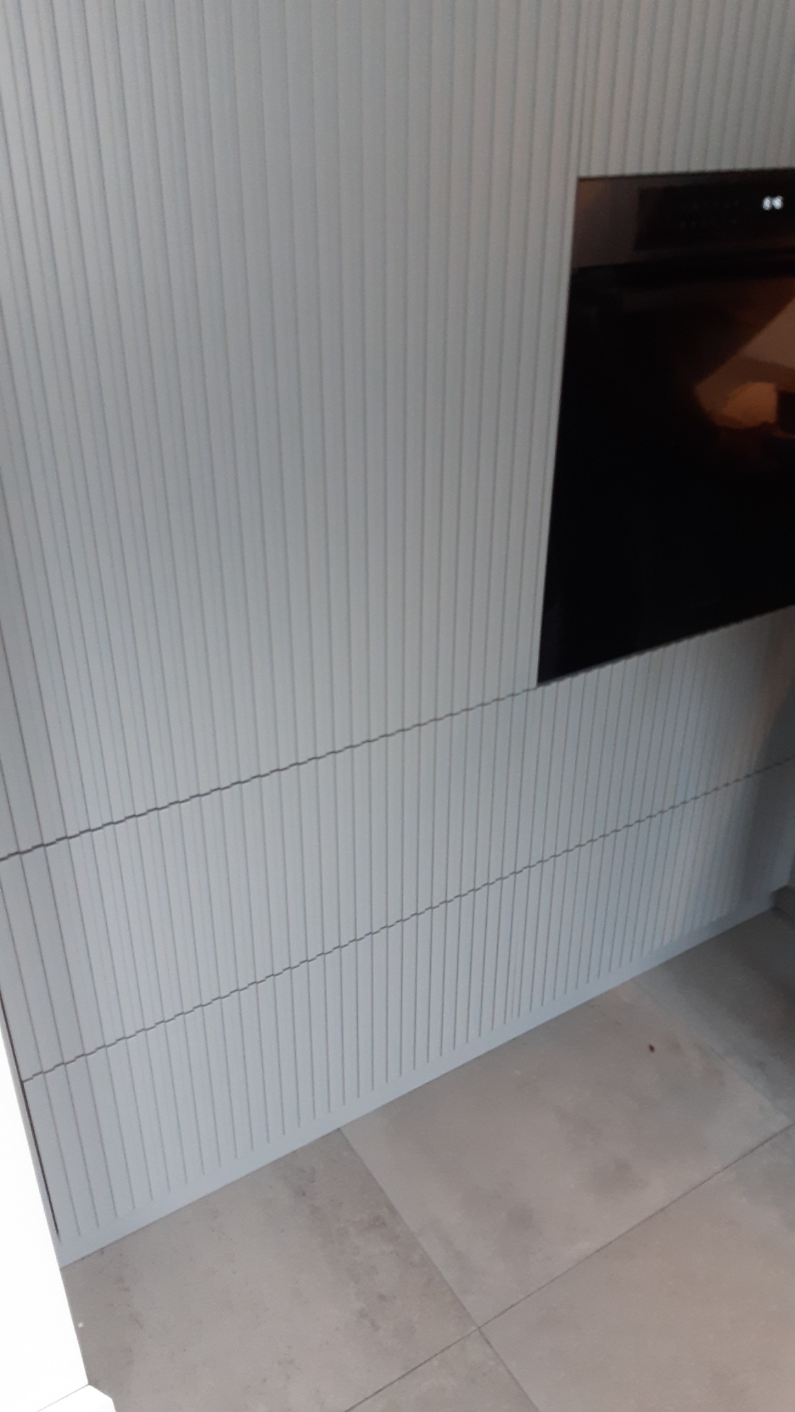 Biała, ryflowana zabudowa kuchenna z wbudowanym piekarnikiem, widoczna z boku. Minimalistyczny design, jasne płytki podłogowe. Ujęcie z dołu.