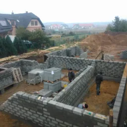 Budowa fundamentów domu z bloczków betonowych, widoczni pracownicy murujący ściany, palety z bloczkami na placu budowy, w tle zabudowa jednorodzinna i zalesiony teren.