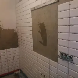 Układanie białych, prostokątnych płytek na ścianie w łazience, widoczny fragment ściany z nałożonym klejem i wystającymi przewodami elektrycznymi, drabina w rogu.