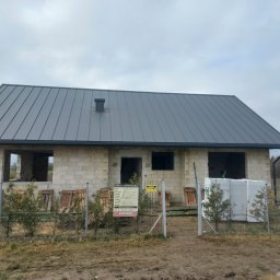 Usługi Blacharsko-Dekarskie SZWAGIER-DACH - Surowy budynek jednorodzinny z szarą blachą na dachu, na działce z ogrodzeniem z siatki, z widoczną tablicą informacyjną budowy i zielonymi sadzonkami iglaków.