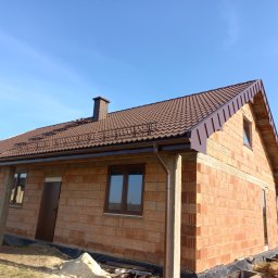Usługi Blacharsko-Dekarskie SZWAGIER-DACH - Dom w stanie surowym otwartym z brązowym dachem z blachodachówki, widoczne obróbki blacharskie i komin, na pierwszym planie materiały budowlane.