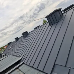 Nowoczesny, grafitowy dach z blachy na rąbek stojący, z widocznymi kominami wentylacyjnymi, na tle pochmurnego nieba z widocznym w oddali żurawiem budowlanym.