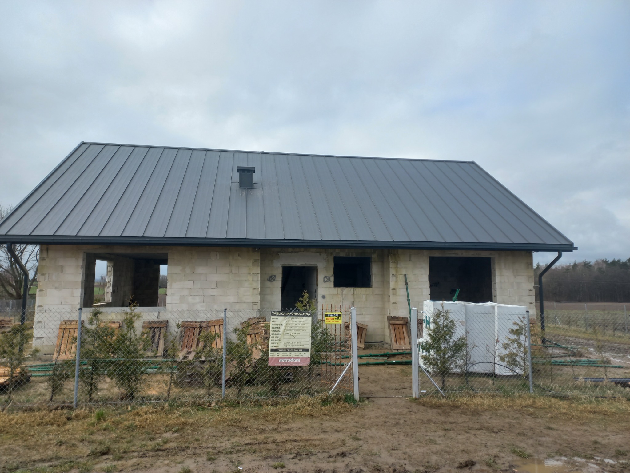 Surowy budynek jednorodzinny z szarą blachą na dachu, na działce z ogrodzeniem z siatki, z widoczną tablicą informacyjną budowy i zielonymi sadzonkami iglaków.