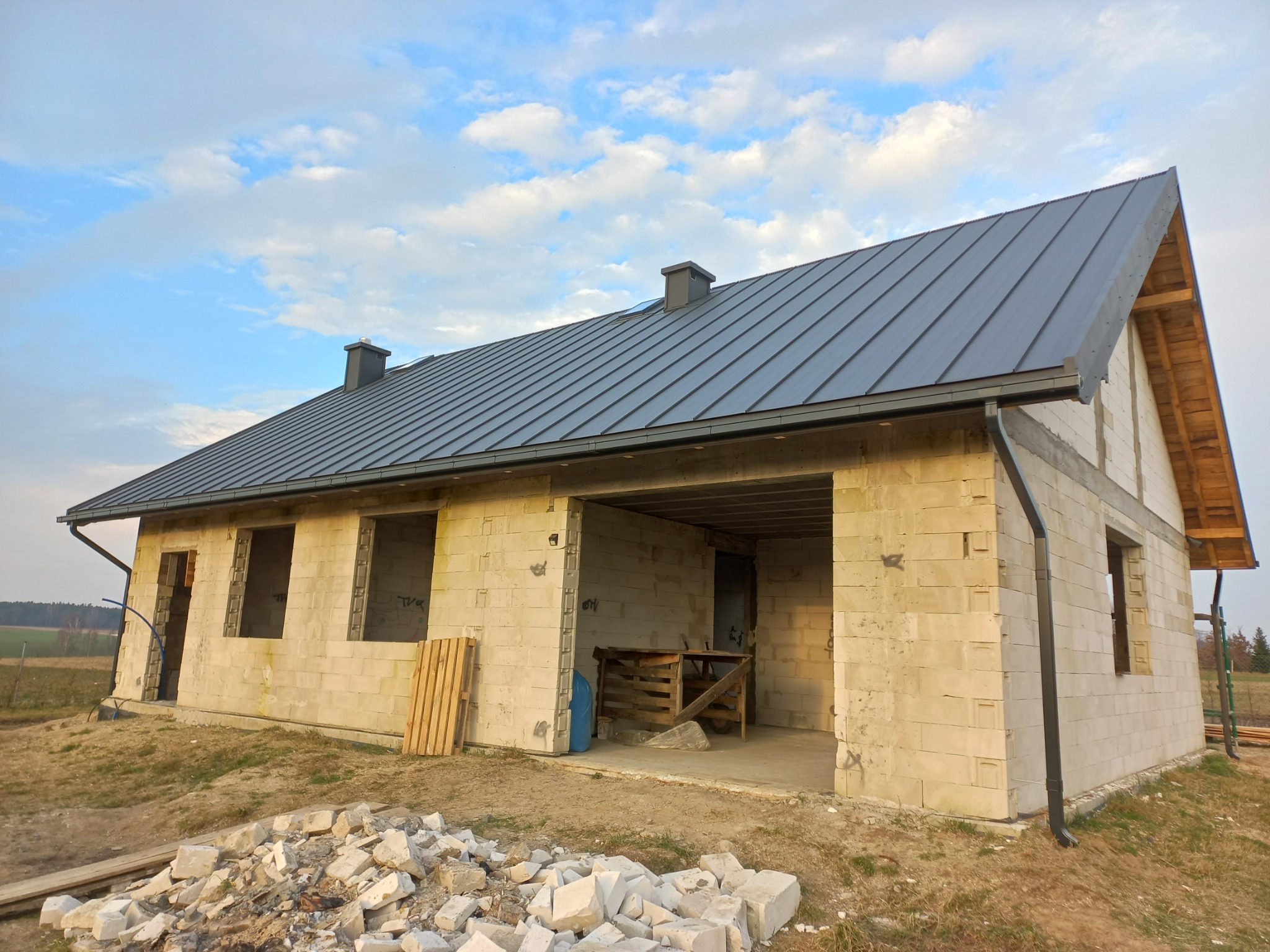 Surowy budynek jednorodzinny z szarą blachą na dachu, widoczne kominy, rynny, niezabudowane okna, palety i gruz budowlany przed budynkiem, błękitne niebo w tle.