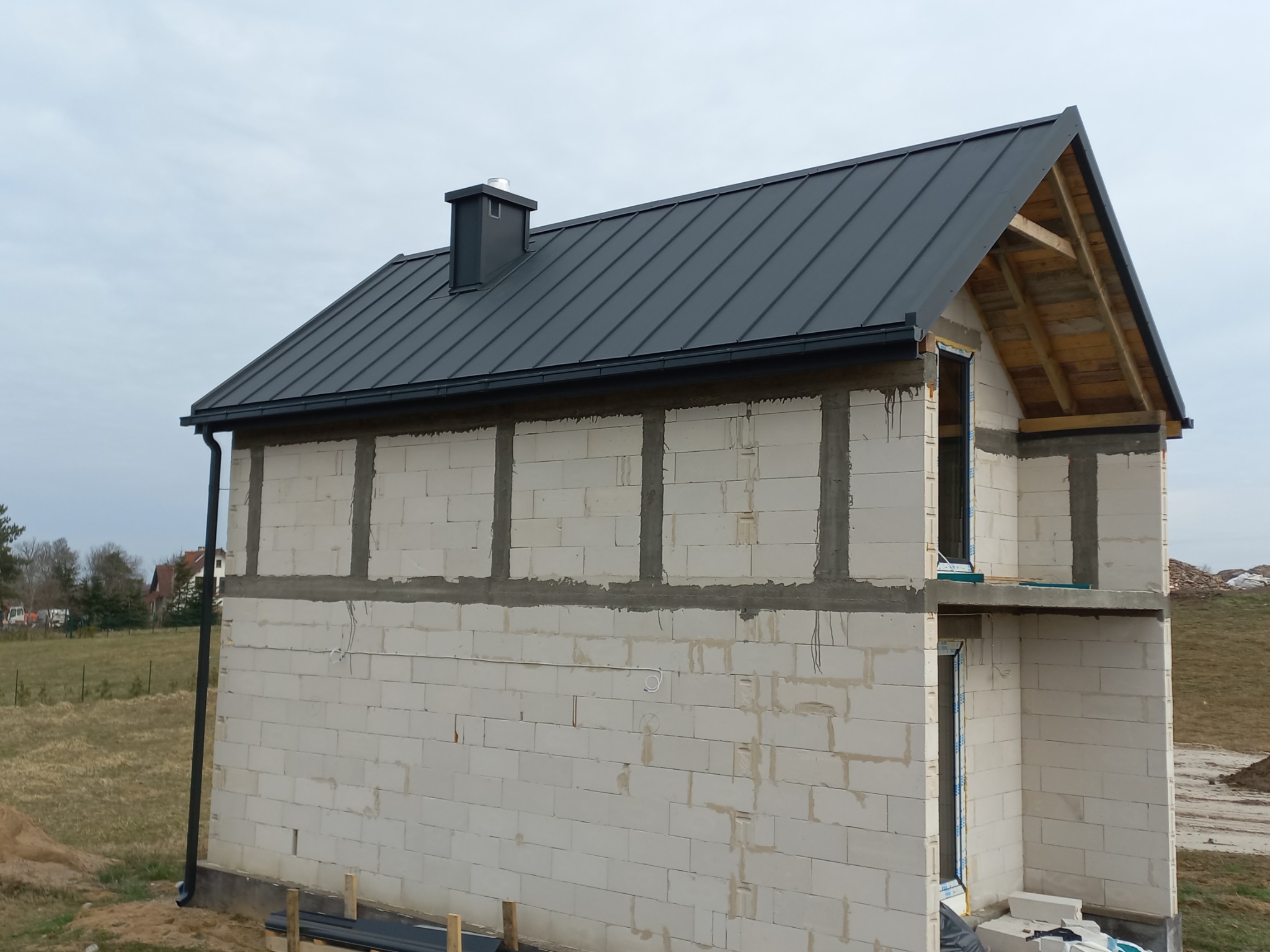 Dom w trakcie budowy z zamontowanym, grafitowym dachem z blachy na rąbek stojący i rynnami w tym samym kolorze, widoczne białe ściany z betonu komórkowego i elementy więźby dachowej.