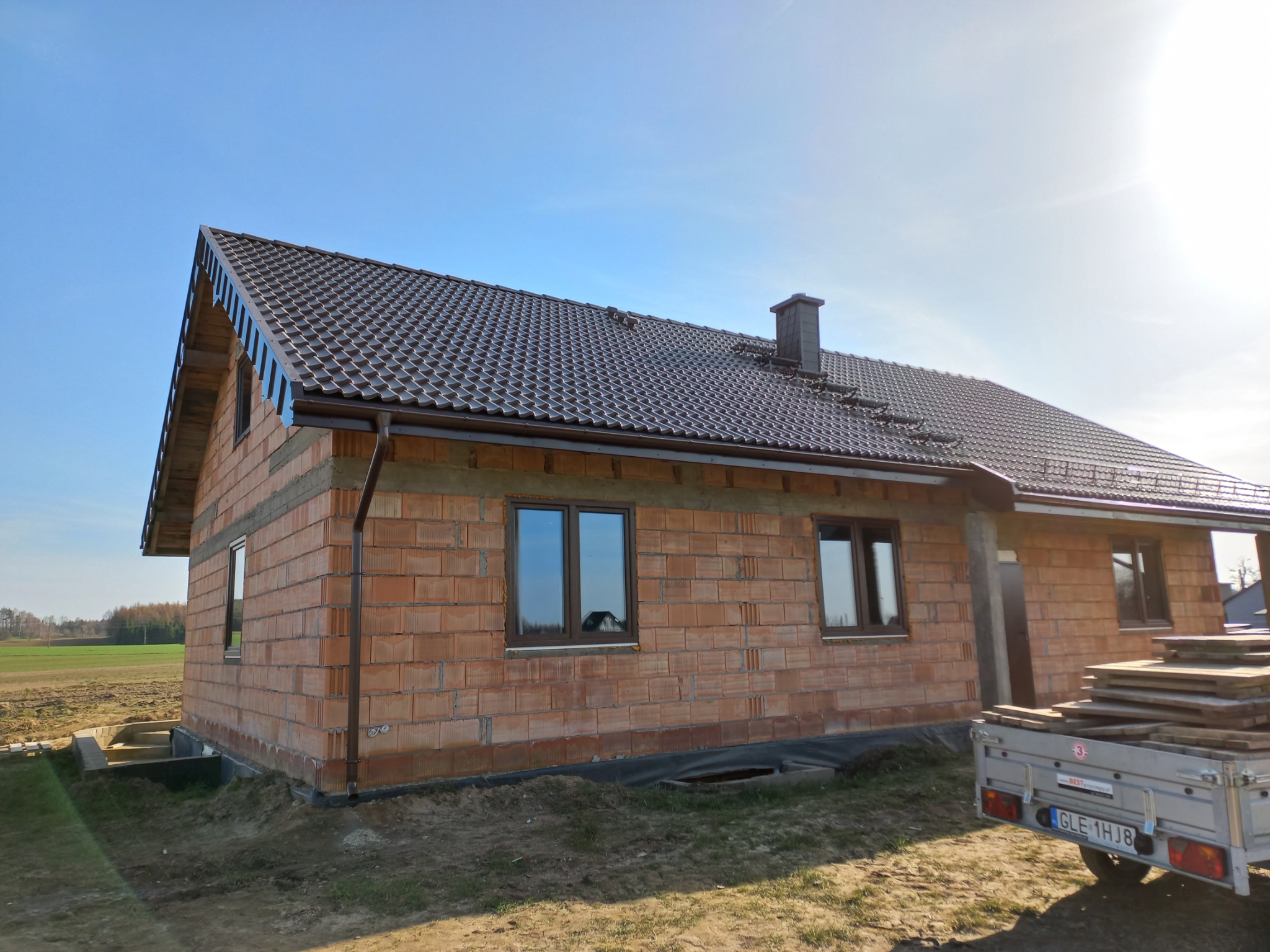 Niedokończony dom jednorodzinny z brązową dachówką i rynnami, widoczne ściany z czerwonej cegły, przyczepka z paletami na pierwszym planie, błękitne niebo w tle.