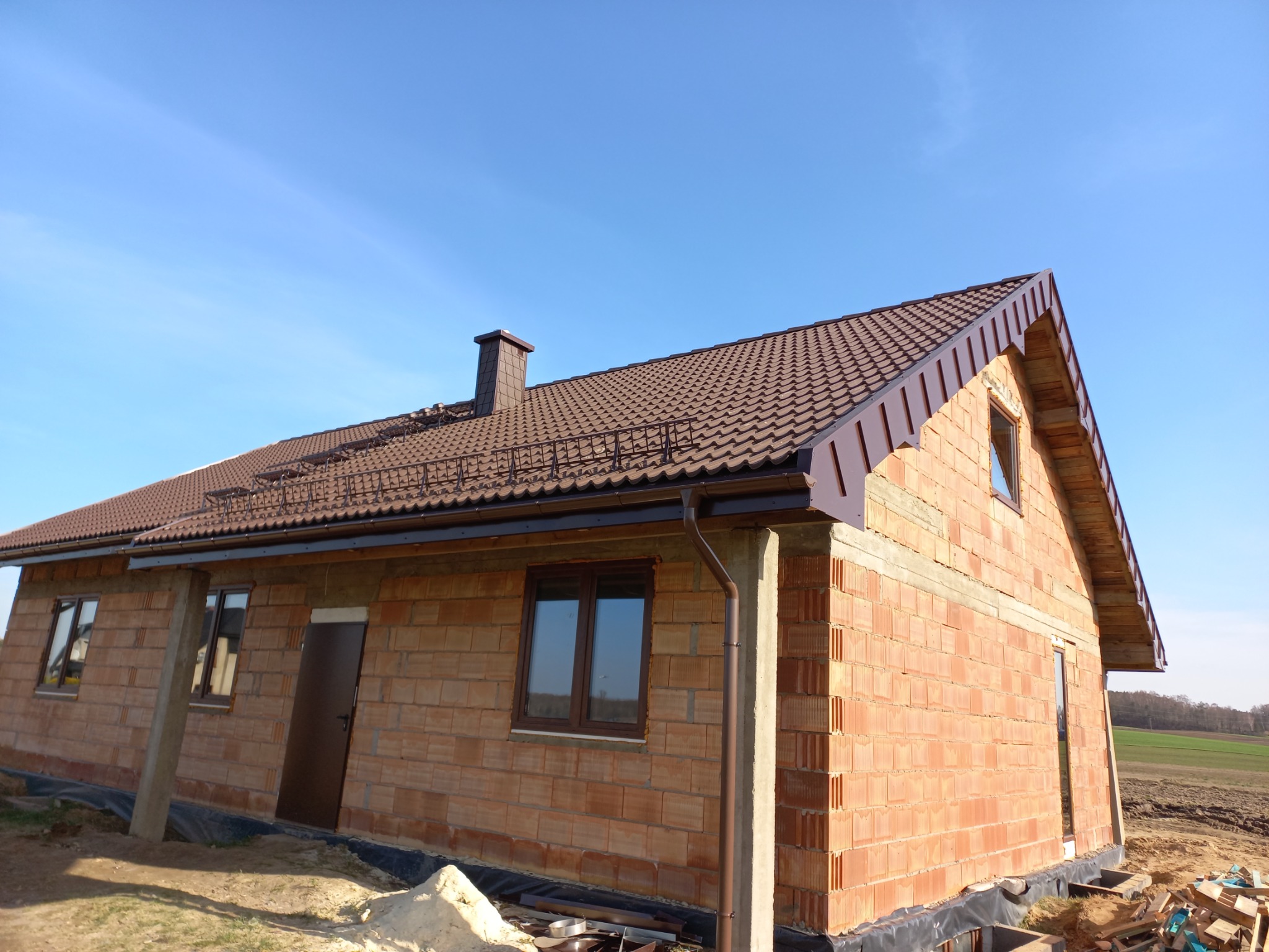 Dom w stanie surowym otwartym z brązowym dachem z blachodachówki, widoczne obróbki blacharskie i komin, na pierwszym planie materiały budowlane.