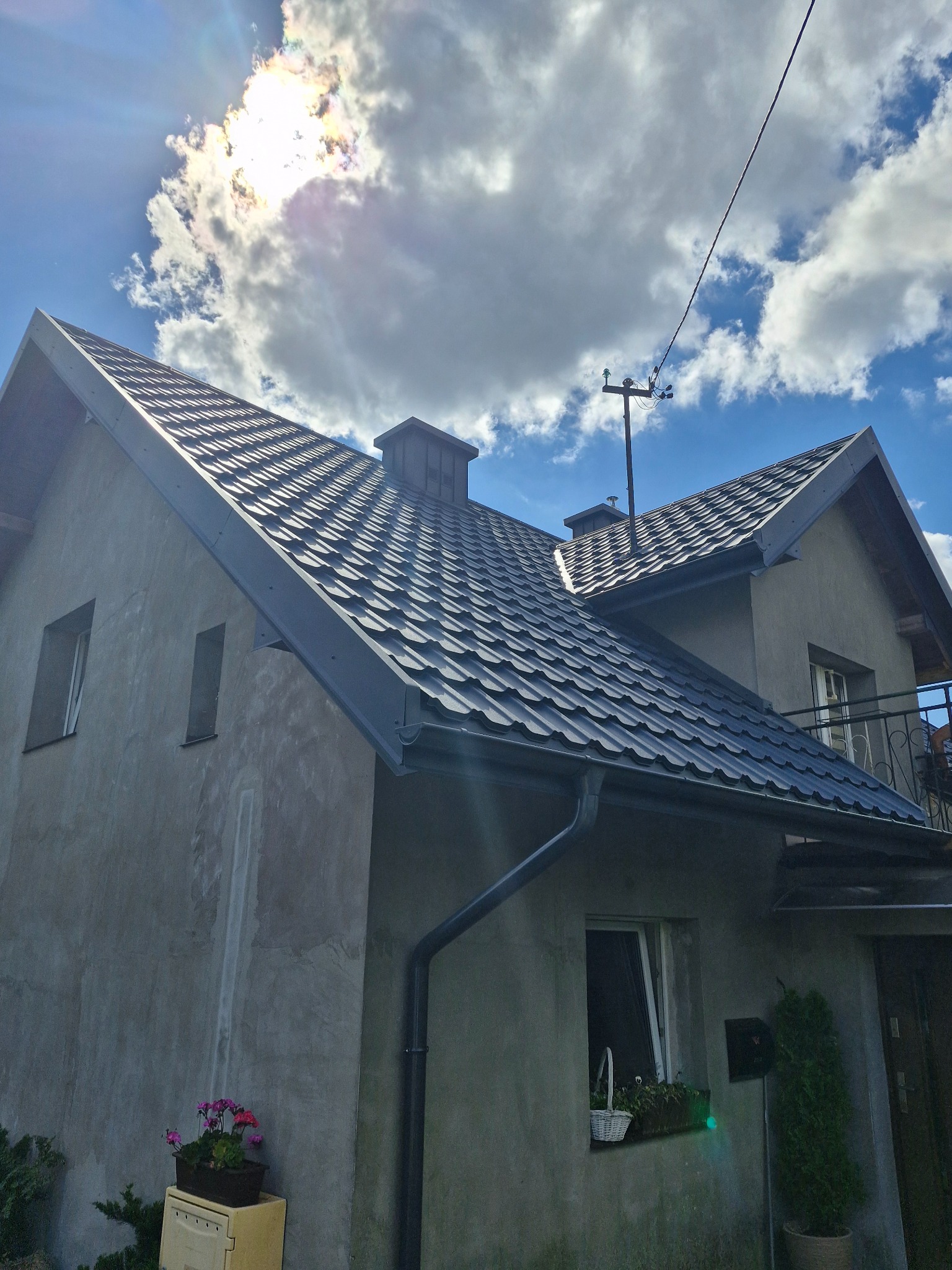 Dom z nowym, grafitowym dachem z blachodachówki modułowej na tle błękitnego nieba z chmurami, widoczny komin i rynna.