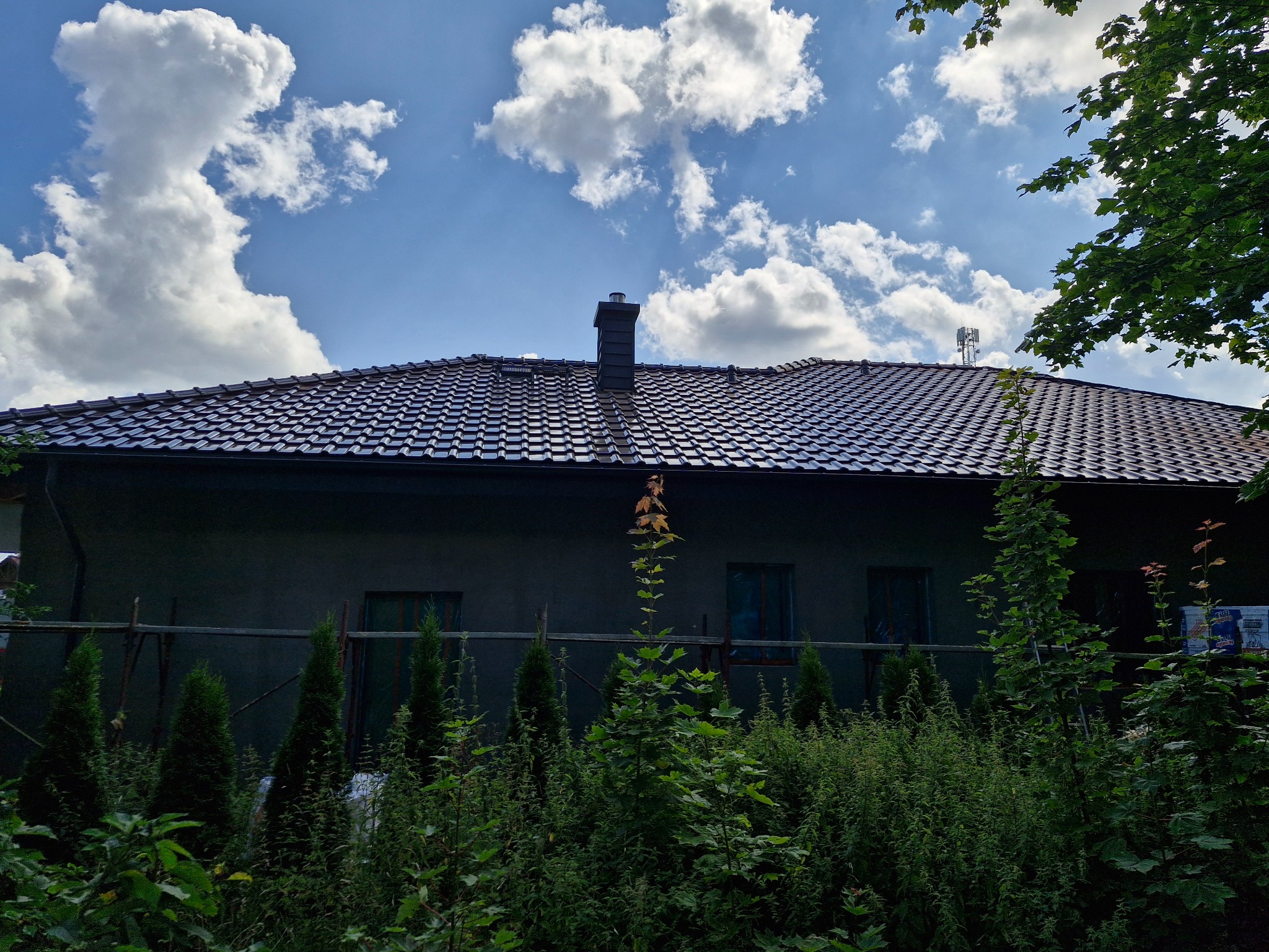 Dom z ciemną dachówką ceramiczną w słoneczny dzień, widoczne rusztowanie i bujna zieleń w ogrodzie.