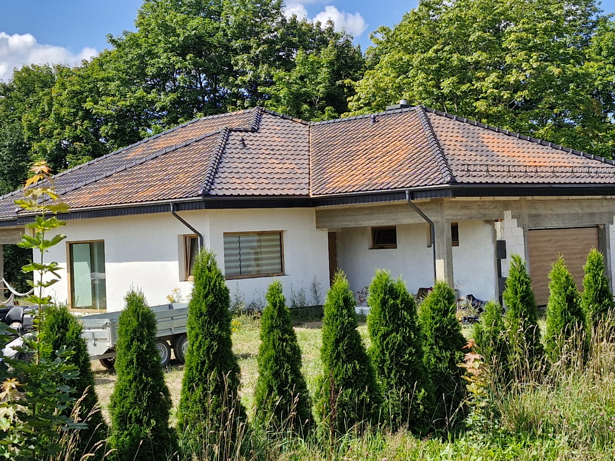 Dom jednorodzinny z brązową dachówką, częściowo pokrytą mchem, z widocznymi rynnami i elementami obróbki blacharskiej, otoczony zielenią i młodymi tujami.