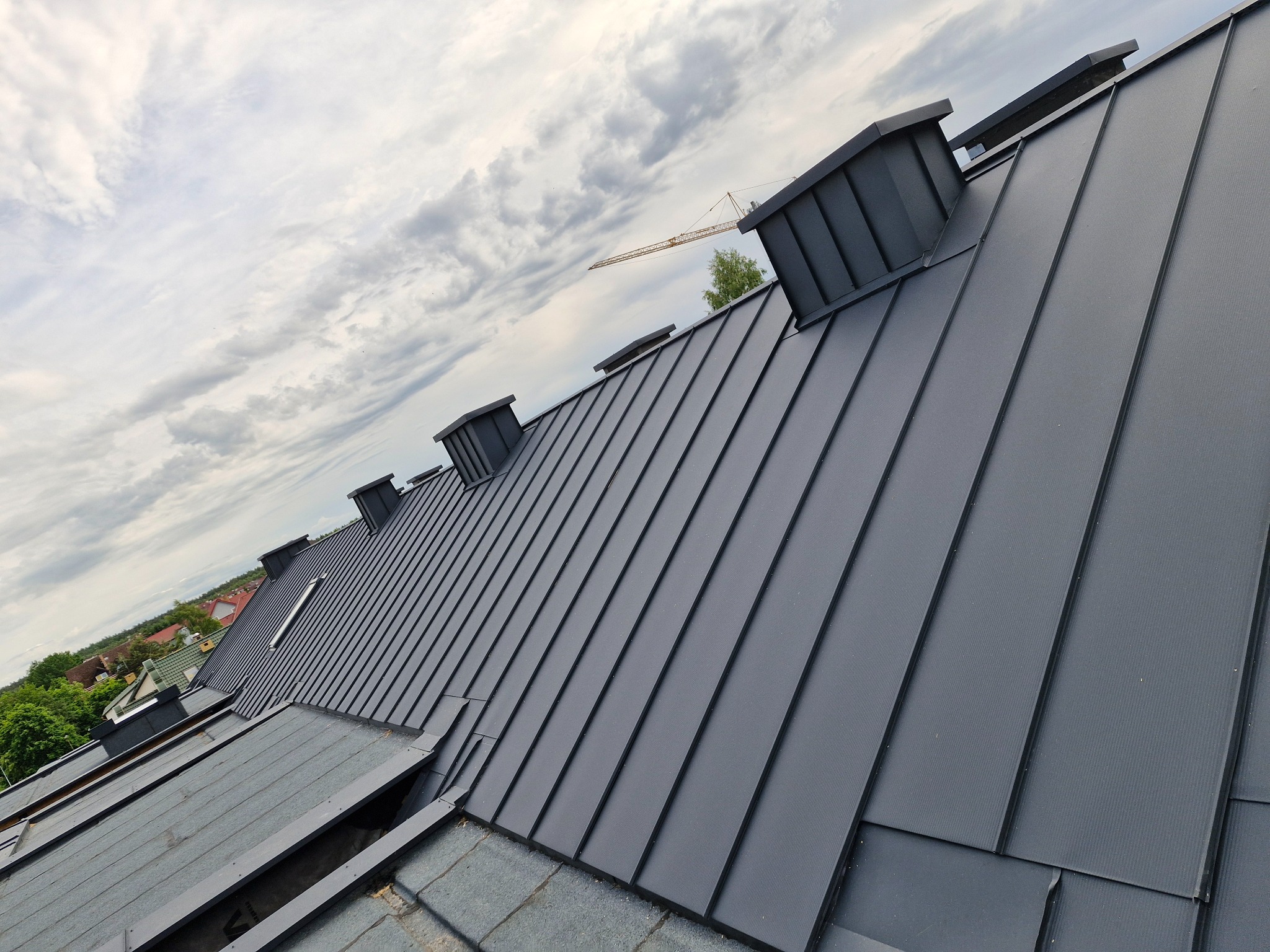 Nowoczesny, grafitowy dach z blachy na rąbek stojący, z widocznymi kominami wentylacyjnymi, na tle pochmurnego nieba z widocznym w oddali żurawiem budowlanym.