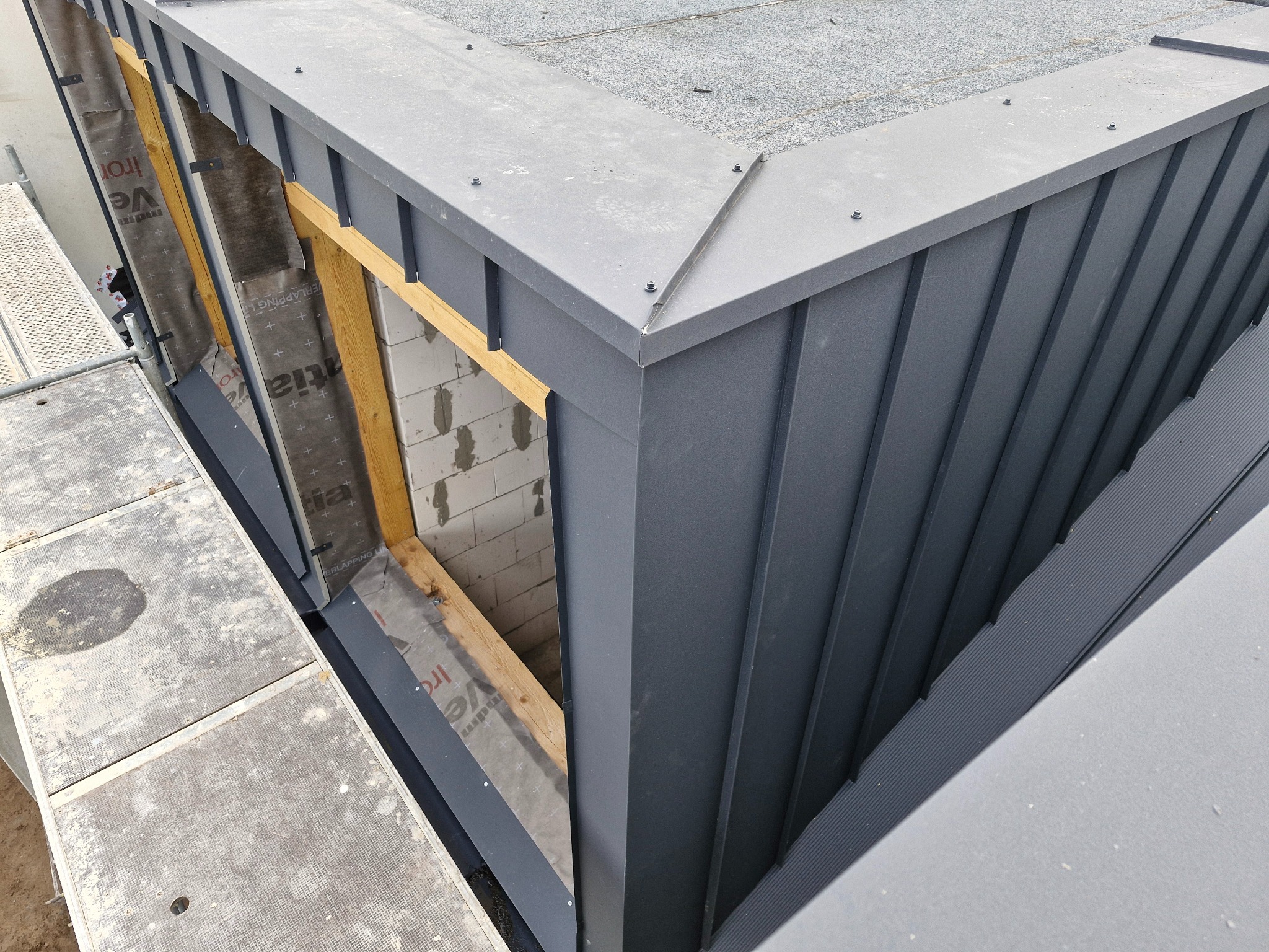 Kąt dachu z nowym, szarym pokryciem blacharskim, widoczny fragment więźby z membraną, ujęcie z góry z rusztowania.