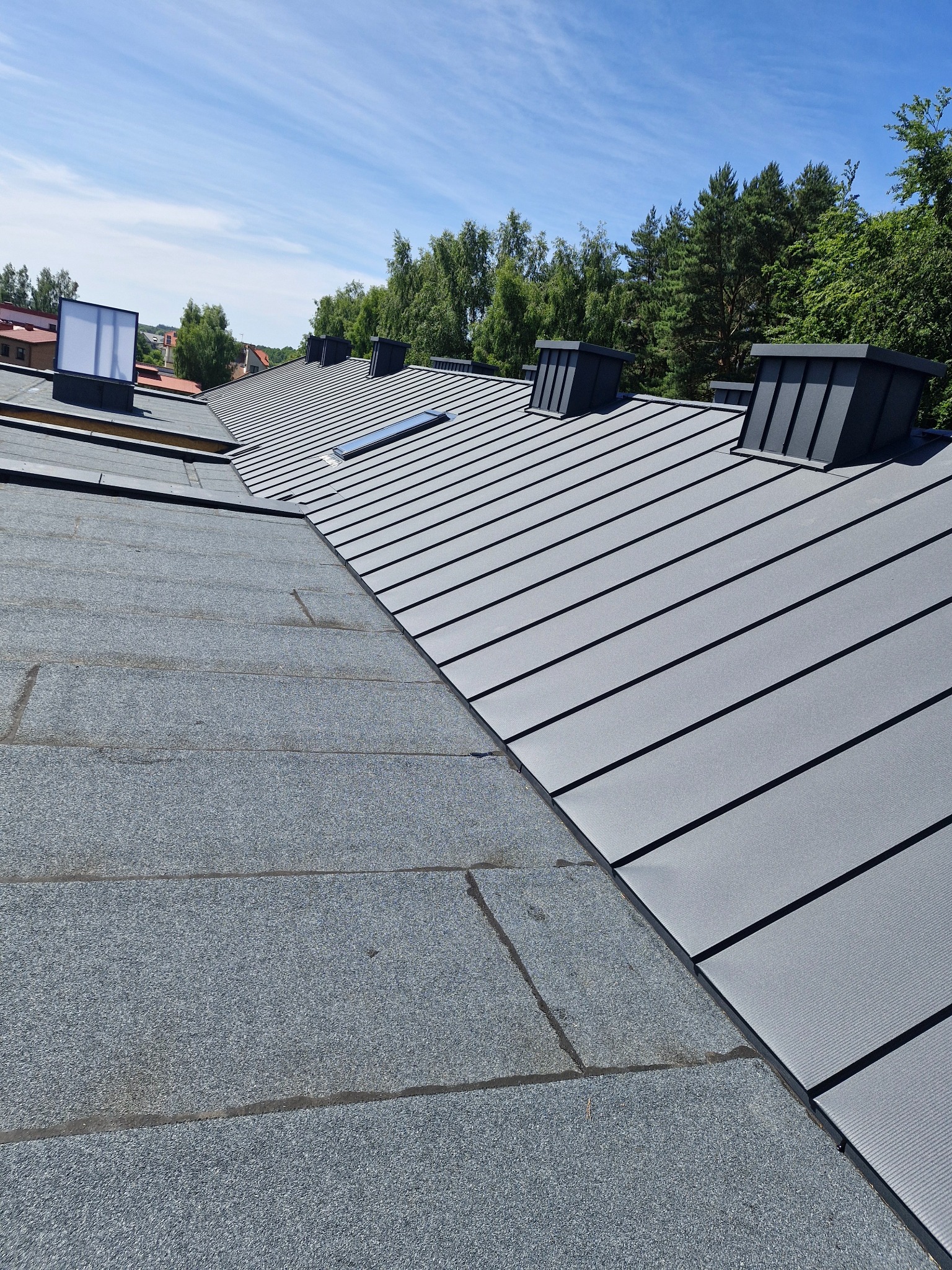 Fragment dachu z widocznym połączeniem dwóch różnych pokryć: papy bitumicznej i blachy na rąbek stojący, z kominkami wentylacyjnymi i oknem dachowym.